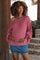 woman wearing a pink off-the-shoulder maternity sweater and a denim skirt, gently cradling her baby bump