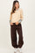 woman wearing a beige ribbed maternity top with patterned sleeves and brown maternity jogger pants, smiling and standing