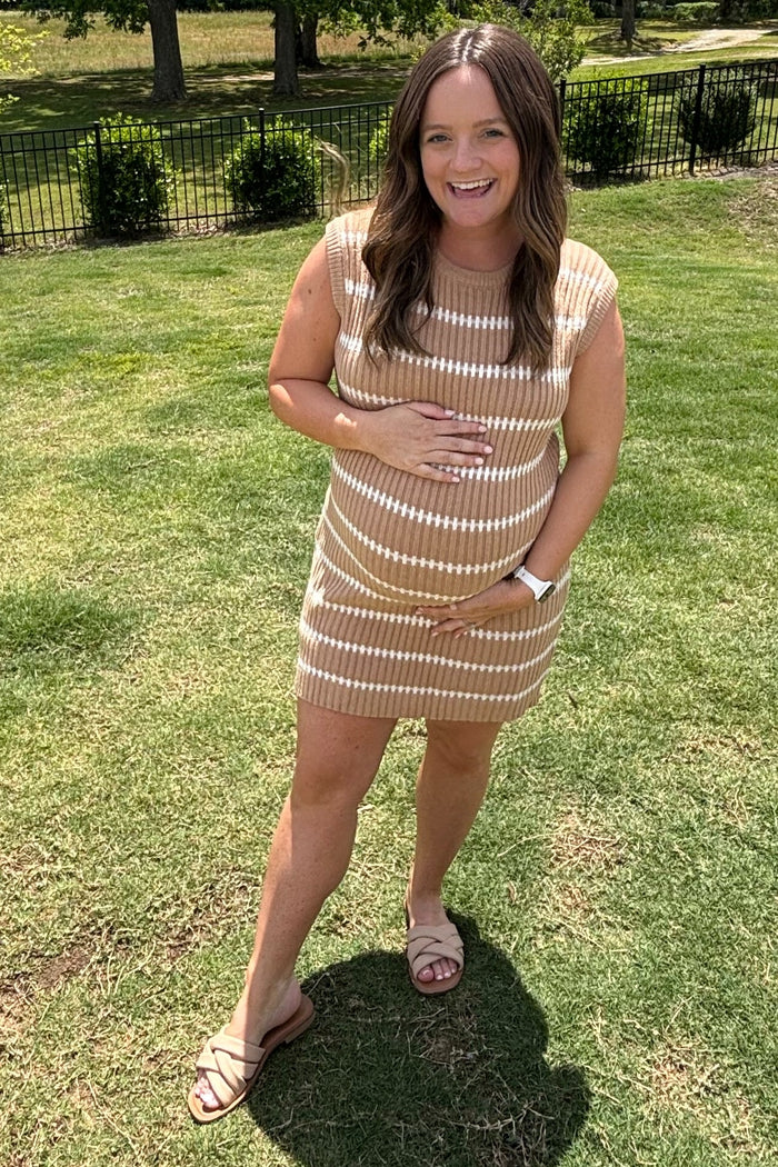 woman wearing a striped beige maternity dress, gently holding her baby bump while standing outdoors
