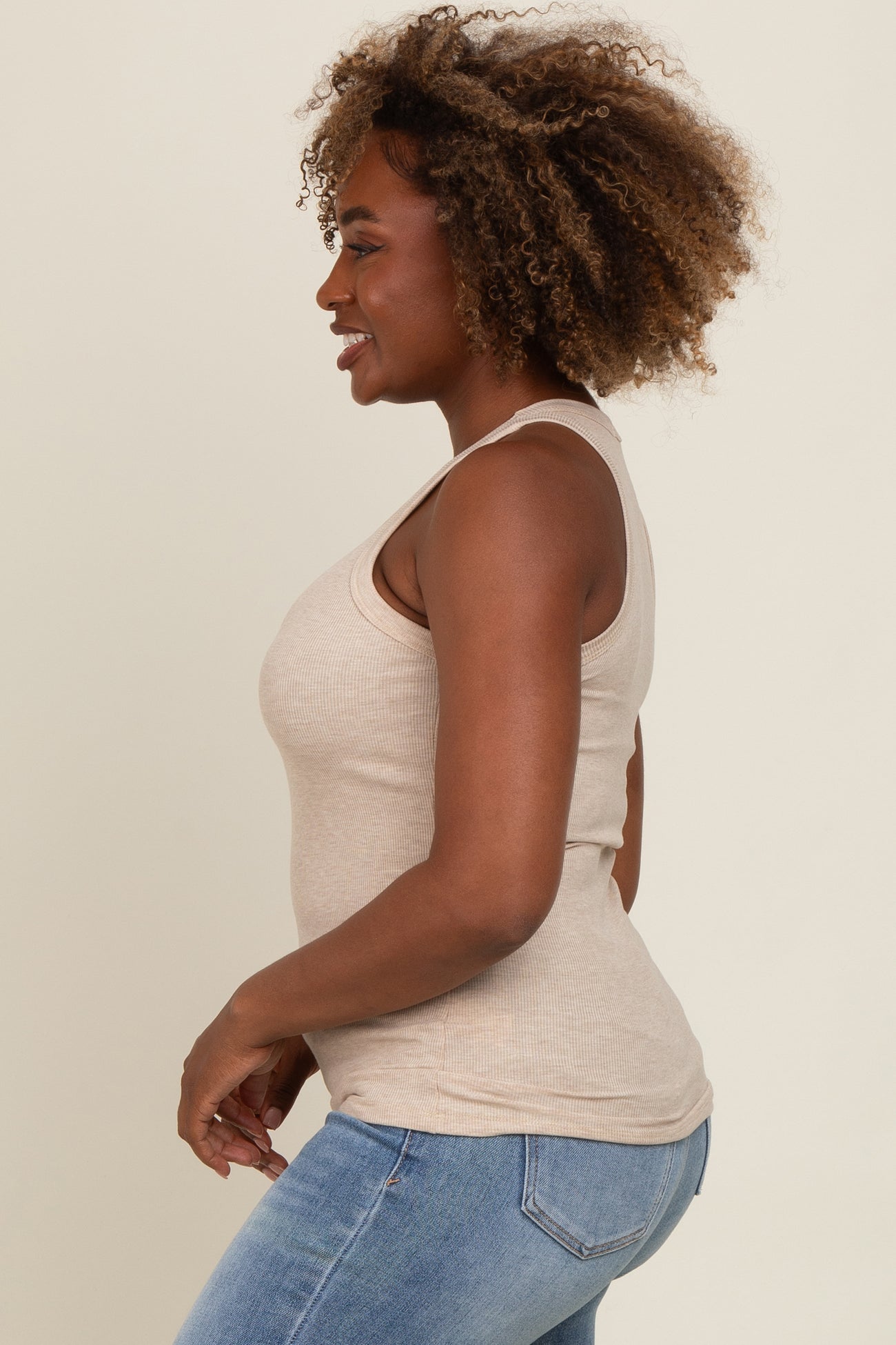 woman wearing a beige ribbed tank top and blue jeans, smiling with curly hair styled down