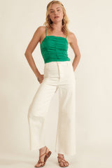 woman wearing a green ruched tank top and white wide-leg pants, posing with one hand on her hip