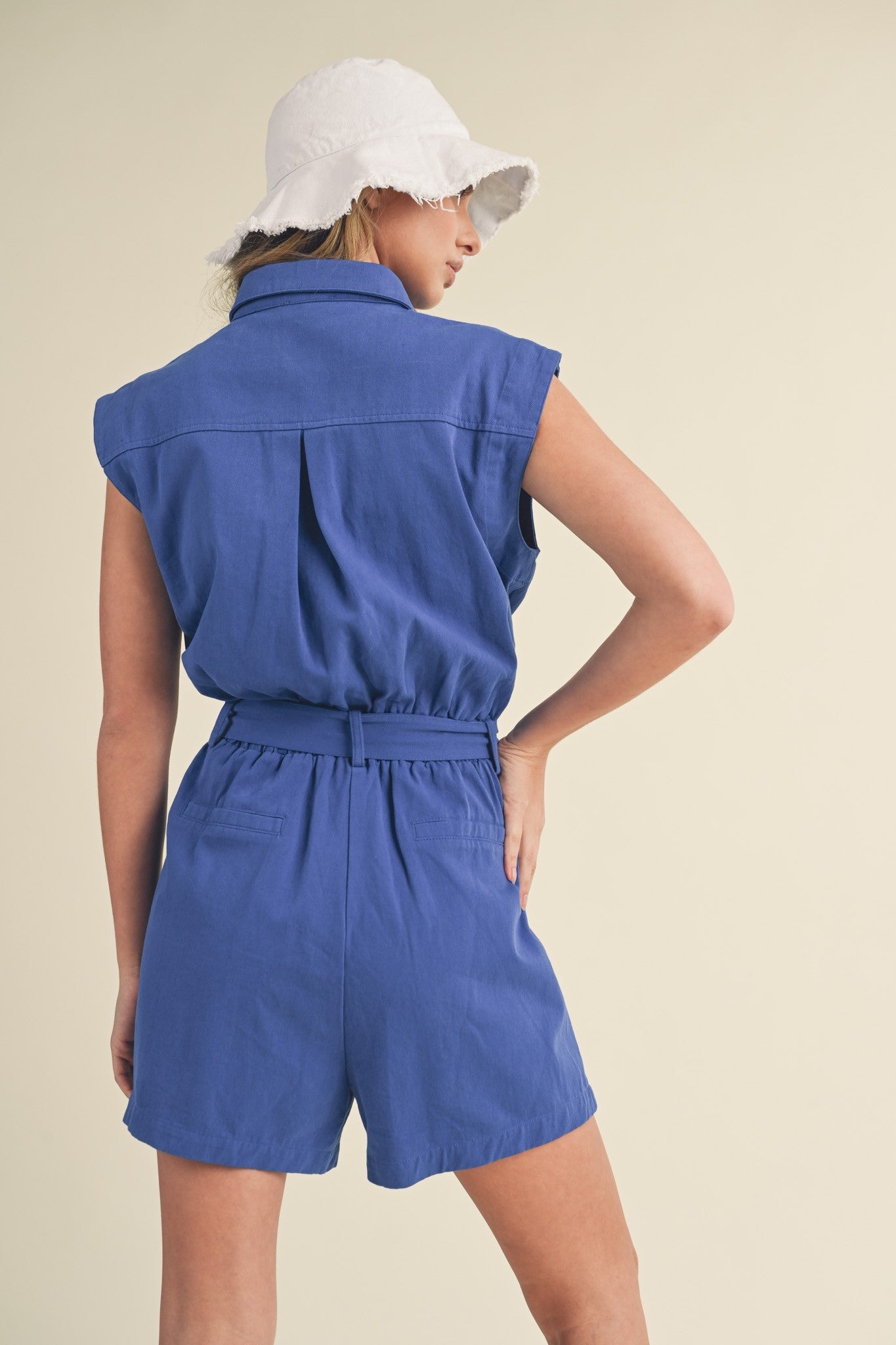 woman wearing a blue sleeveless romper with a belted waist and a white bucket hat, viewed from the back