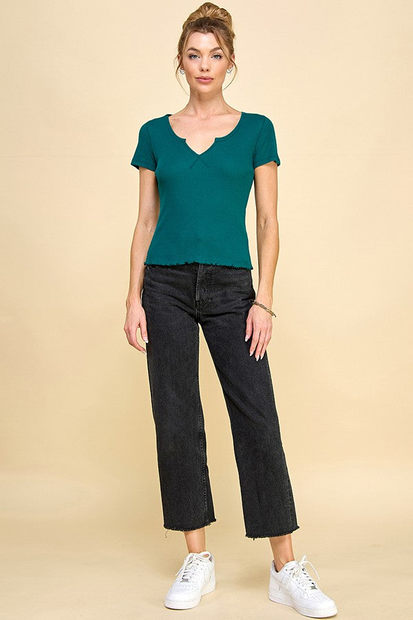 woman wearing a teal short sleeve top with a v-neck and black cropped jeans, standing against a tan background