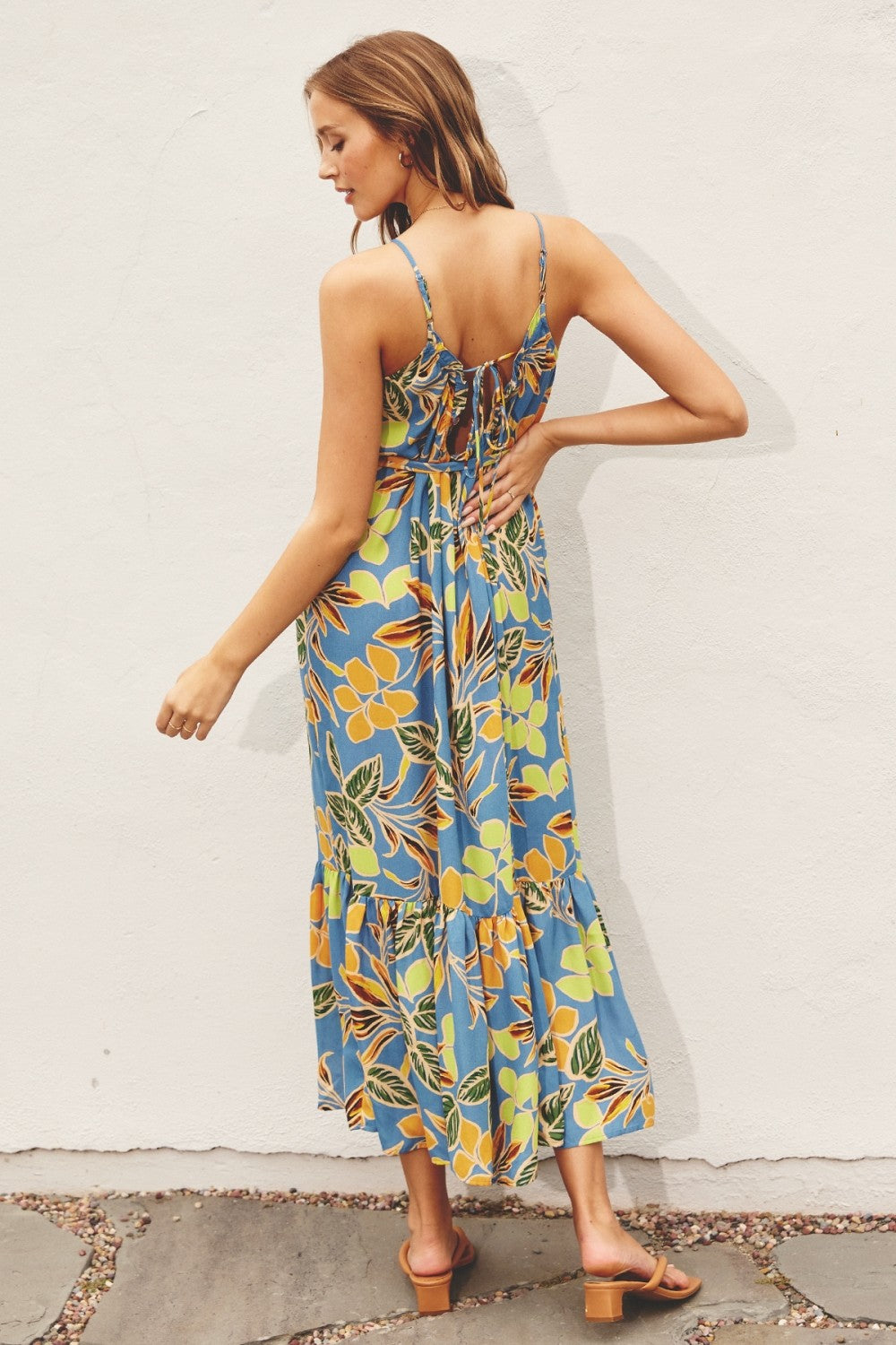 woman wearing a blue floral maxi dress with adjustable straps and a tiered hem, standing against a white wall