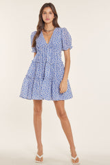 woman wearing a short, blue floral dress with puffed sleeves and a v-neckline, standing against a plain background