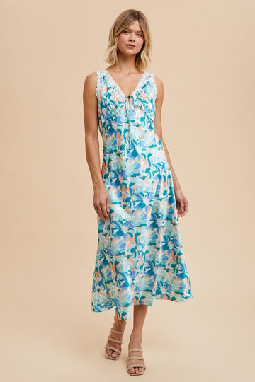 woman wearing a blue floral sleeveless maxi dress with lace detailing and a tie front, styled with neutral sandals