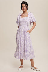 woman wearing a lavender floral print midi dress with puff sleeves and a smocked bodice, standing with one hand in a pocket