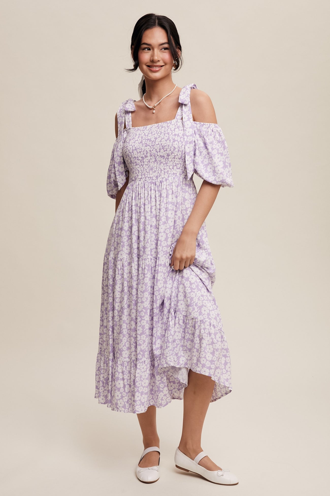 woman wearing a lavender floral off-the-shoulder dress with puff sleeves and fitted bodice, smiling and holding the hem