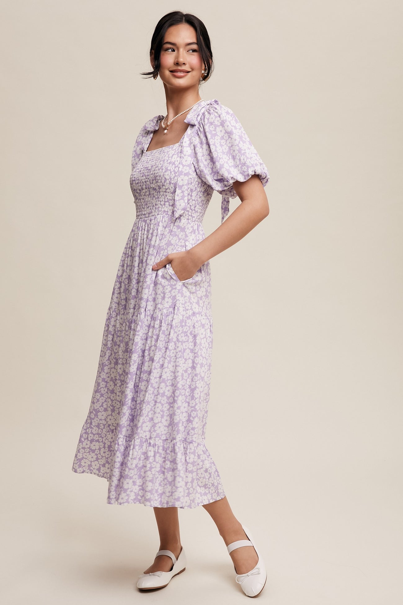 woman wearing a lavender floral midi dress with puff sleeves and pockets, smiling against a neutral background