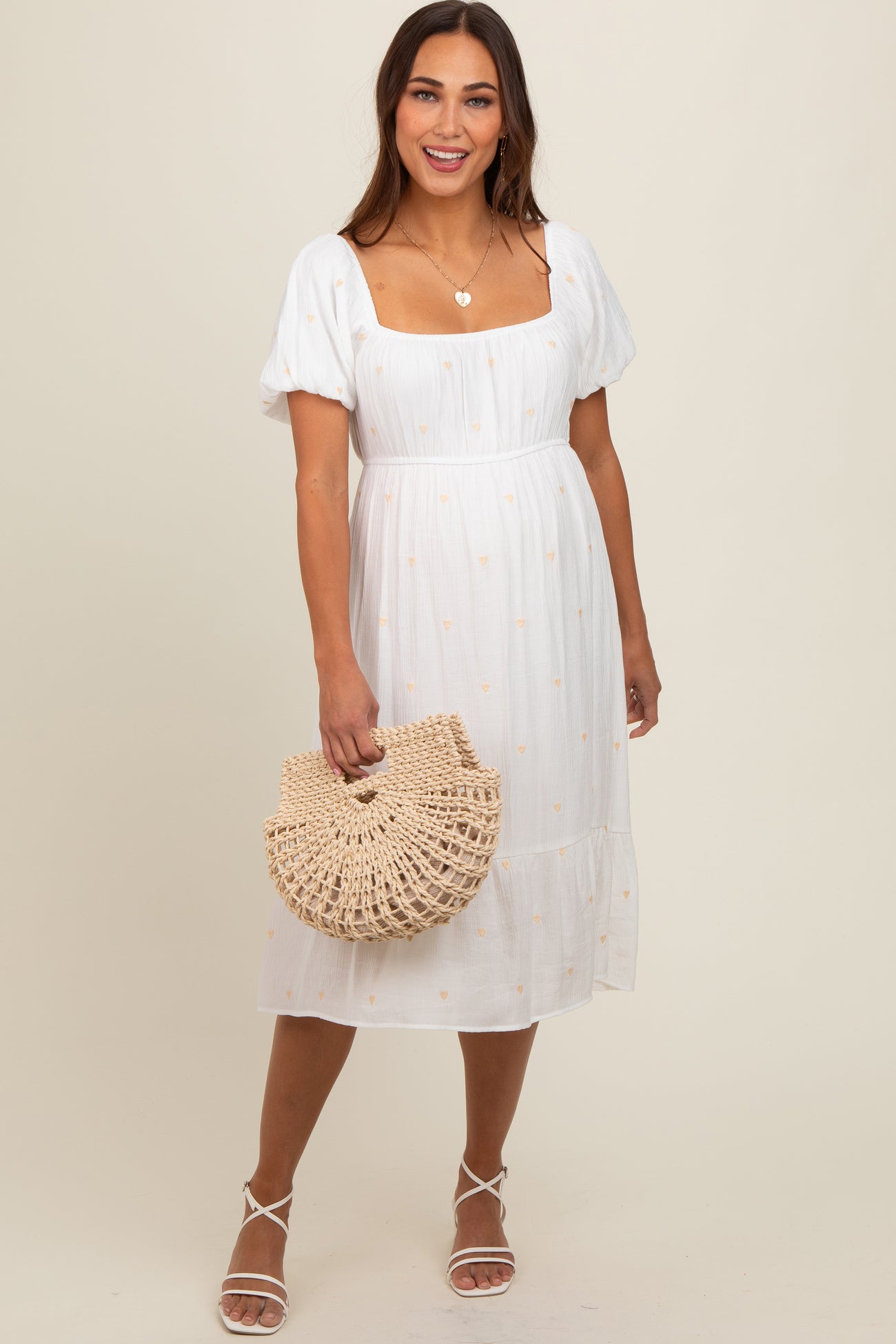 woman wearing a white maternity dress with puff sleeves and embroidered details, holding a woven bag