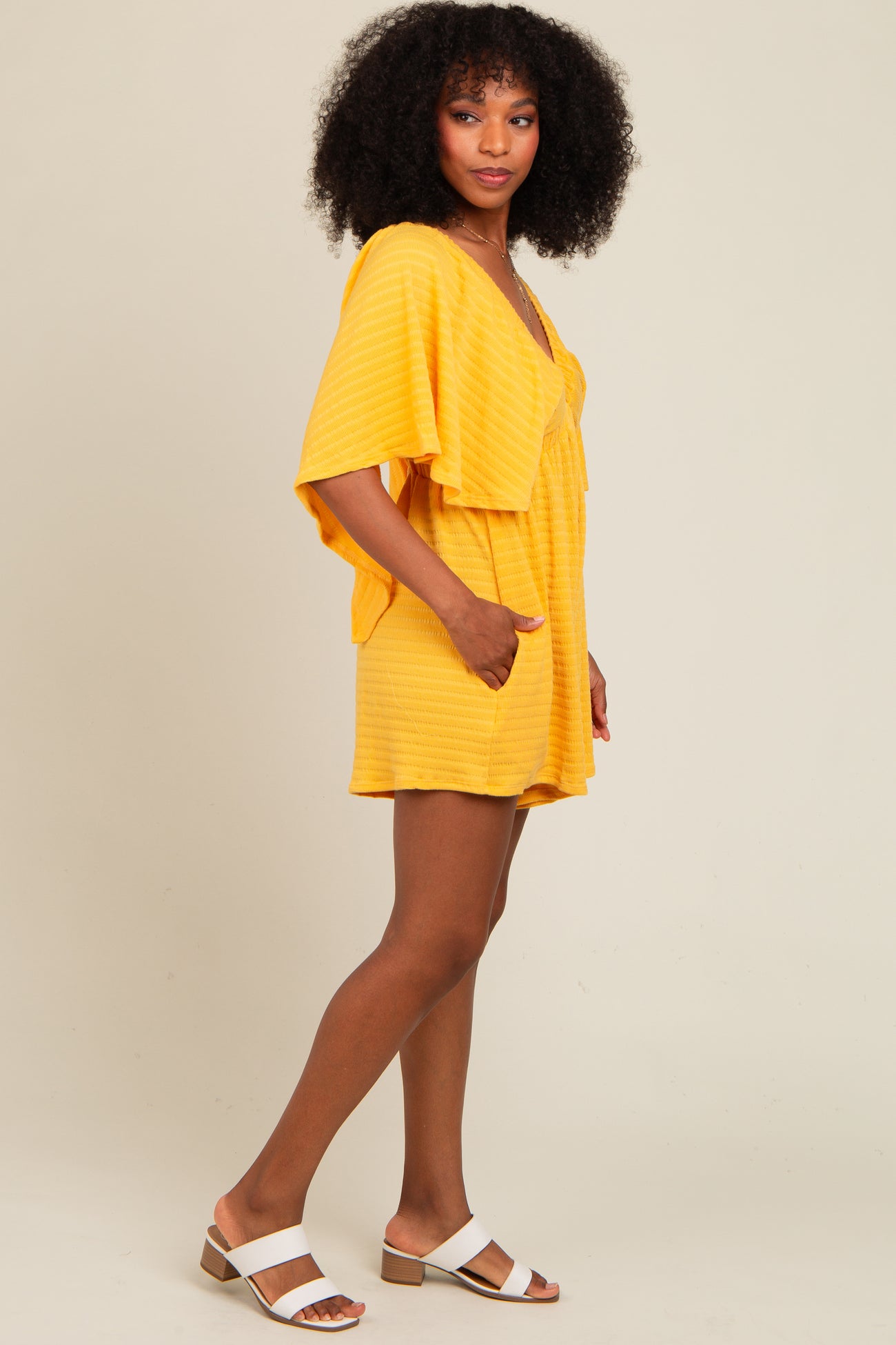 woman wearing a yellow striped short romper with flutter sleeves and side pockets, paired with white sandals