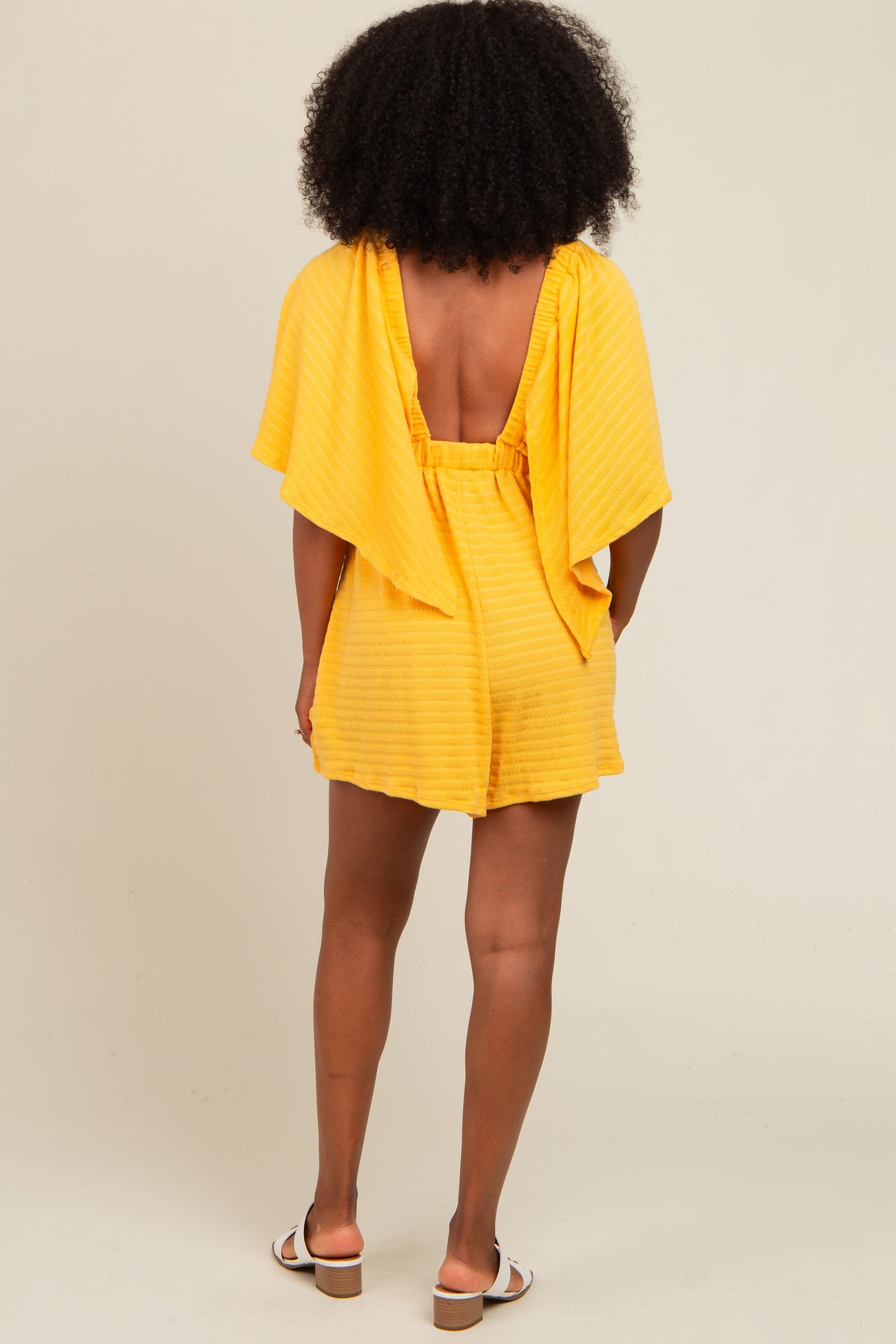 woman wearing a yellow striped romper with a backless design and flutter sleeves, standing in a neutral background