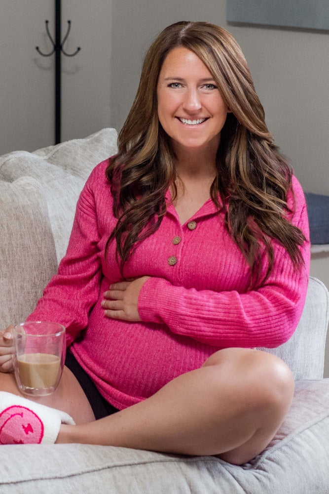 woman wearing a pink ribbed maternity sweater holding a glass of coffee and showing her baby bump while sitting on a couch