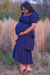 woman wearing a navy blue maternity two-piece outfit with a tiered skirt, holding her baby bump in a grassy field