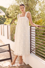 woman wearing a white midi dress with ruffled straps and floral details, standing by a railing outdoors