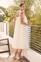 woman wearing a white sleeveless dress with floral appliqués and bow details, standing outdoors near a railing
