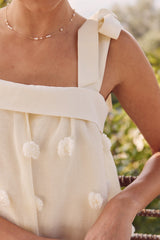 woman wearing a cream sleeveless top with a bow on one strap and textured pom-pom details