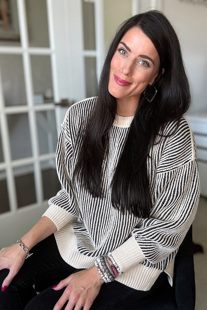 woman wearing a cream and black striped oversized sweater, sitting and smiling, with long dark hair and accessories