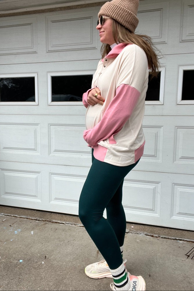 woman wearing a beige and pink maternity pullover with a baby bump, paired with dark green leggings and sunglasses