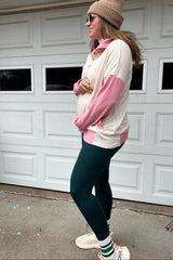 woman wearing a beige and pink maternity pullover with a baby bump, paired with dark green leggings and sunglasses