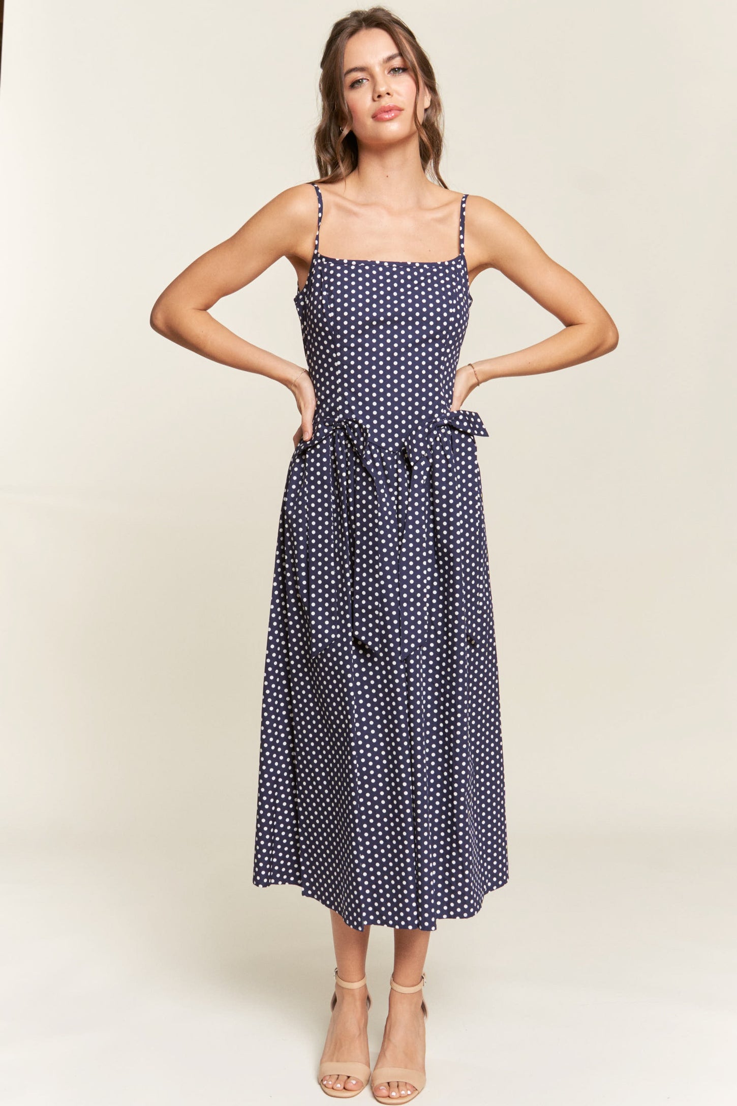 woman wearing a navy blue polka dot midi dress with adjustable straps and a tie belt at the waist