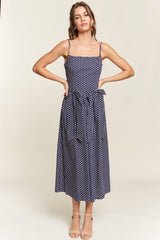 woman wearing a navy blue polka dot midi dress with adjustable straps and a tie belt at the waist