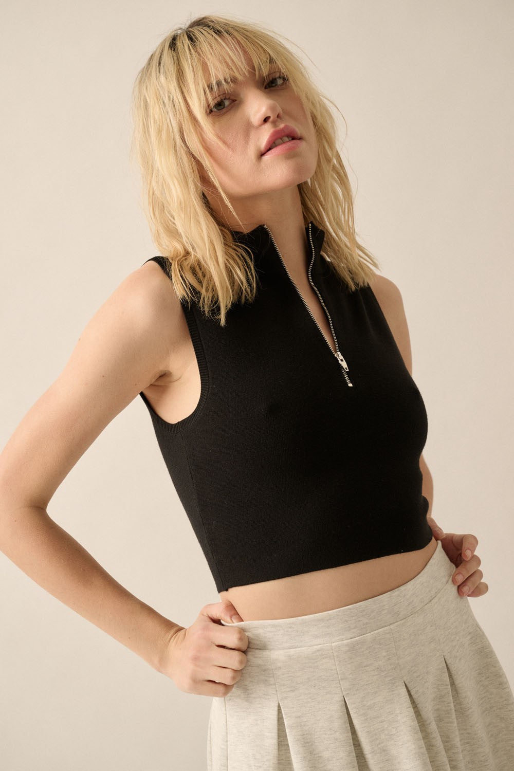 woman wearing a black cropped sleeveless top with a zip front and light gray pleated skirt