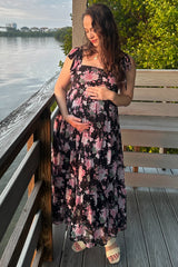 woman wearing a black floral maxi maternity dress and holding her baby bump by the water's edge