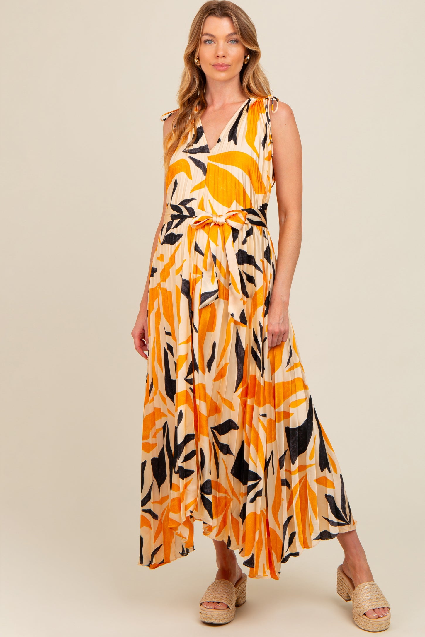 woman wearing a sleeveless yellow and orange patterned maternity maxi dress with a flowy skirt and adjustable shoulder ties, baby bump visible