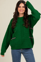 woman wearing a green cable-knit sweater with pink accents and long sleeves, posing with one hand raised
