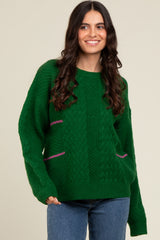 woman wearing a green cable knit sweater with pink detailing and long sleeves, smiling against a neutral backdrop