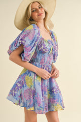 woman wearing a floral purple mini dress with puff sleeves and smocked waist, styled with a wide-brimmed hat