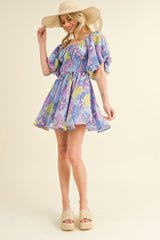 woman wearing a floral off-the-shoulder mini dress in purple and blue, paired with a wide-brimmed hat and platform sandals