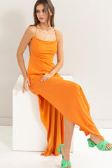 woman wearing an orange spaghetti strap maxi dress and green high heeled sandals, sitting on a white pedestal