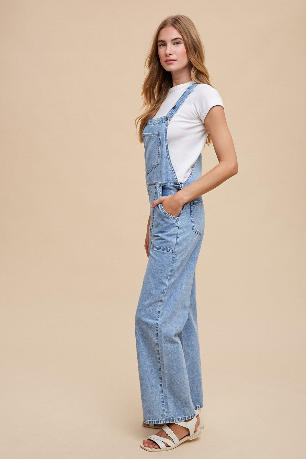 woman wearing a white short sleeve t-shirt and light blue denim overalls, standing against a neutral background