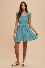 woman wearing a teal smocked mini dress with ruffle sleeves and a tiered skirt, standing against a beige background