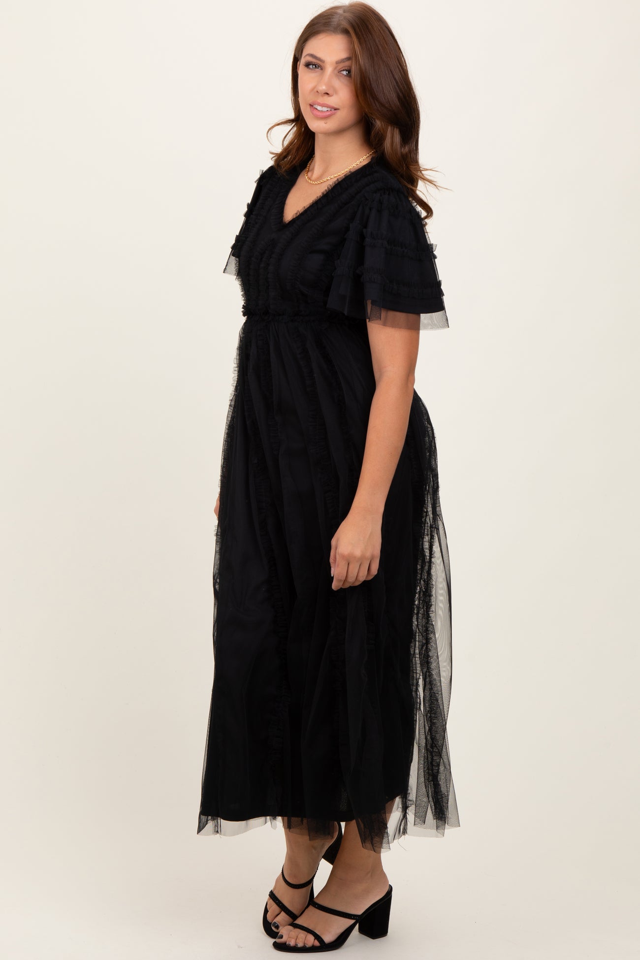 woman wearing a black maxi dress with ruffled sleeves and sheer overlay, standing in a neutral background