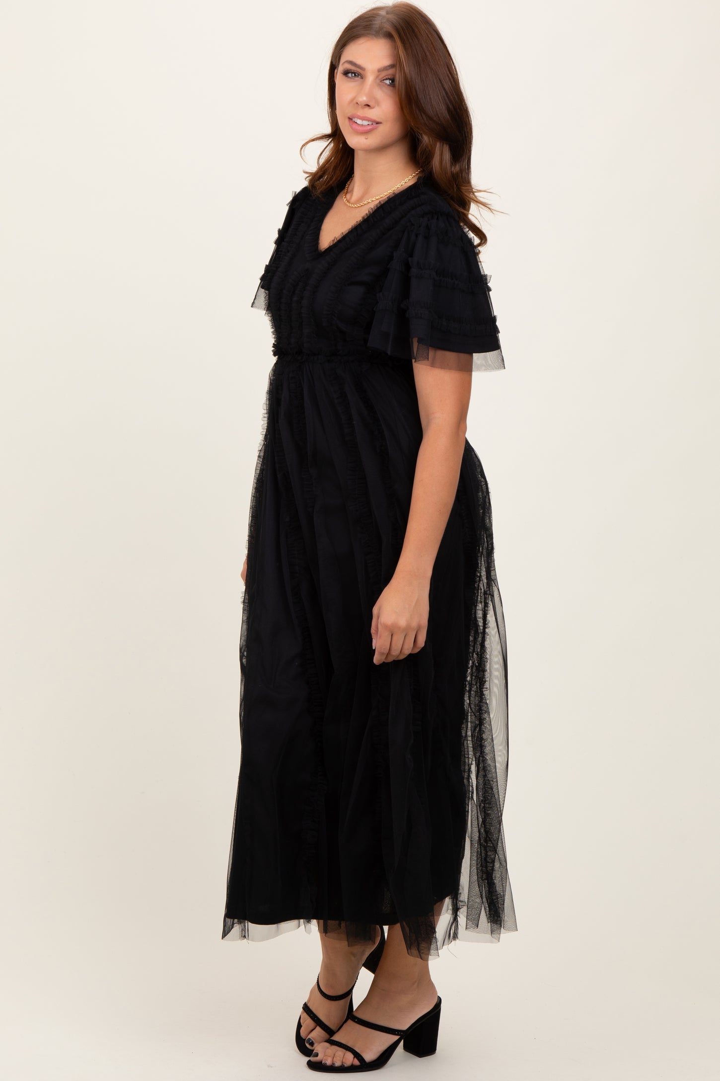 woman wearing a black maxi dress with ruffled sleeves and sheer overlay, standing in a neutral background