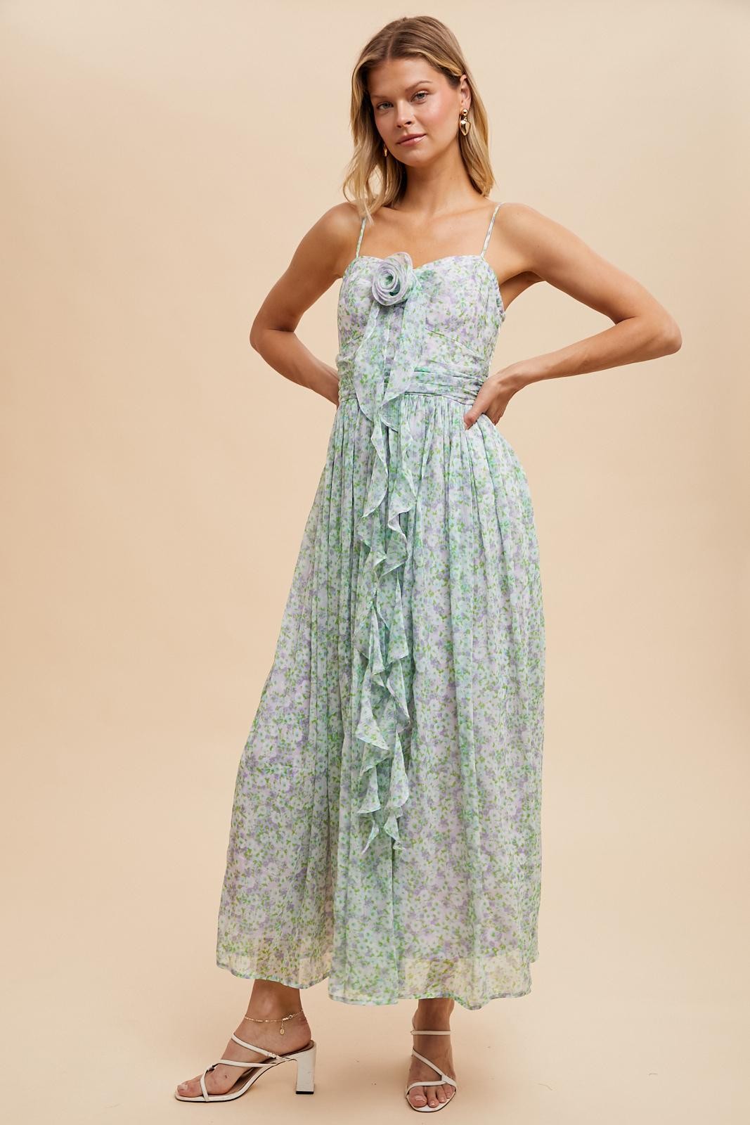 woman wearing a light green floral maxi dress with ruffles and a rose detail, standing with one hand on her hip