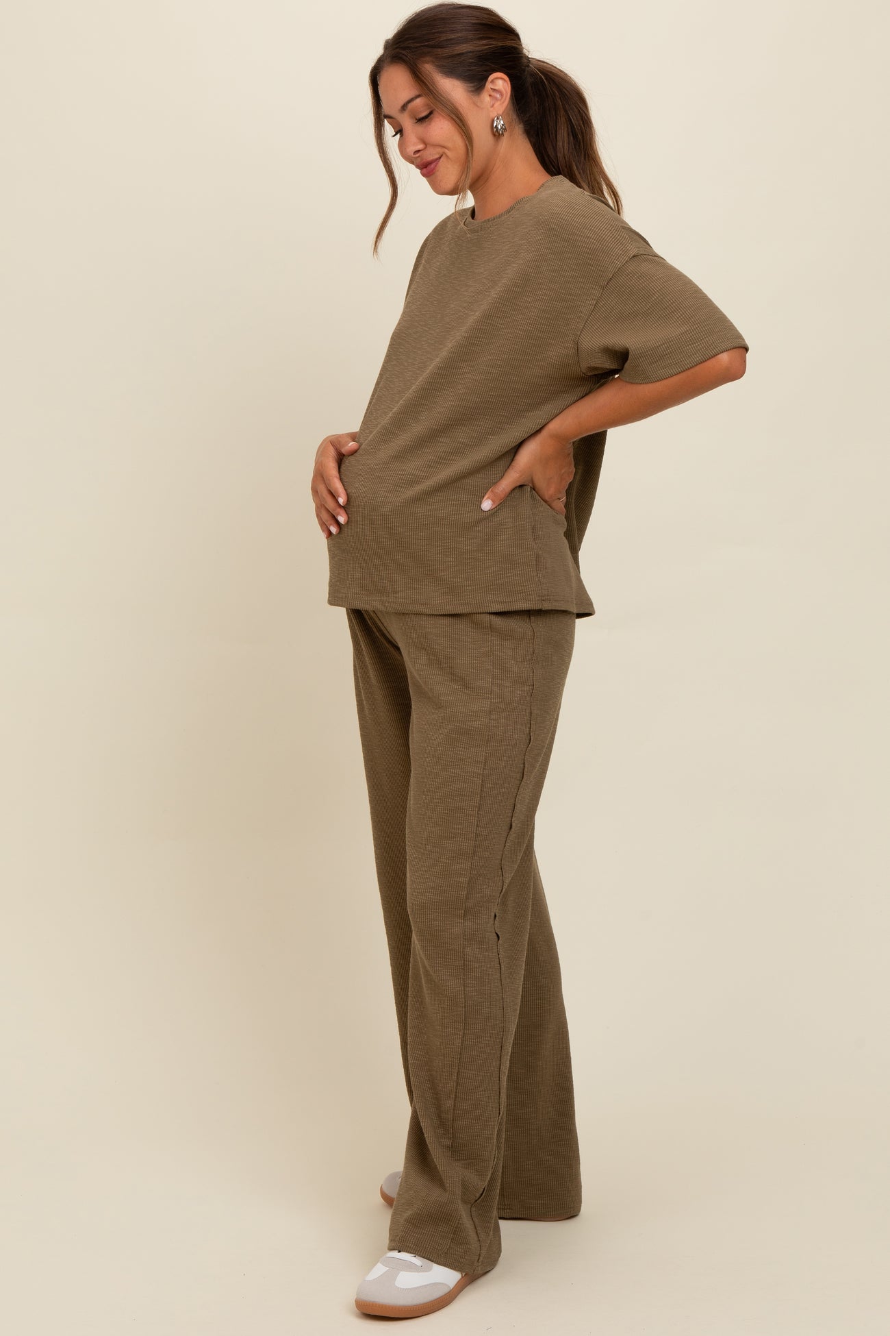 woman wearing a khaki maternity loungewear set with a loose top and wide-leg pants, holding her baby bump