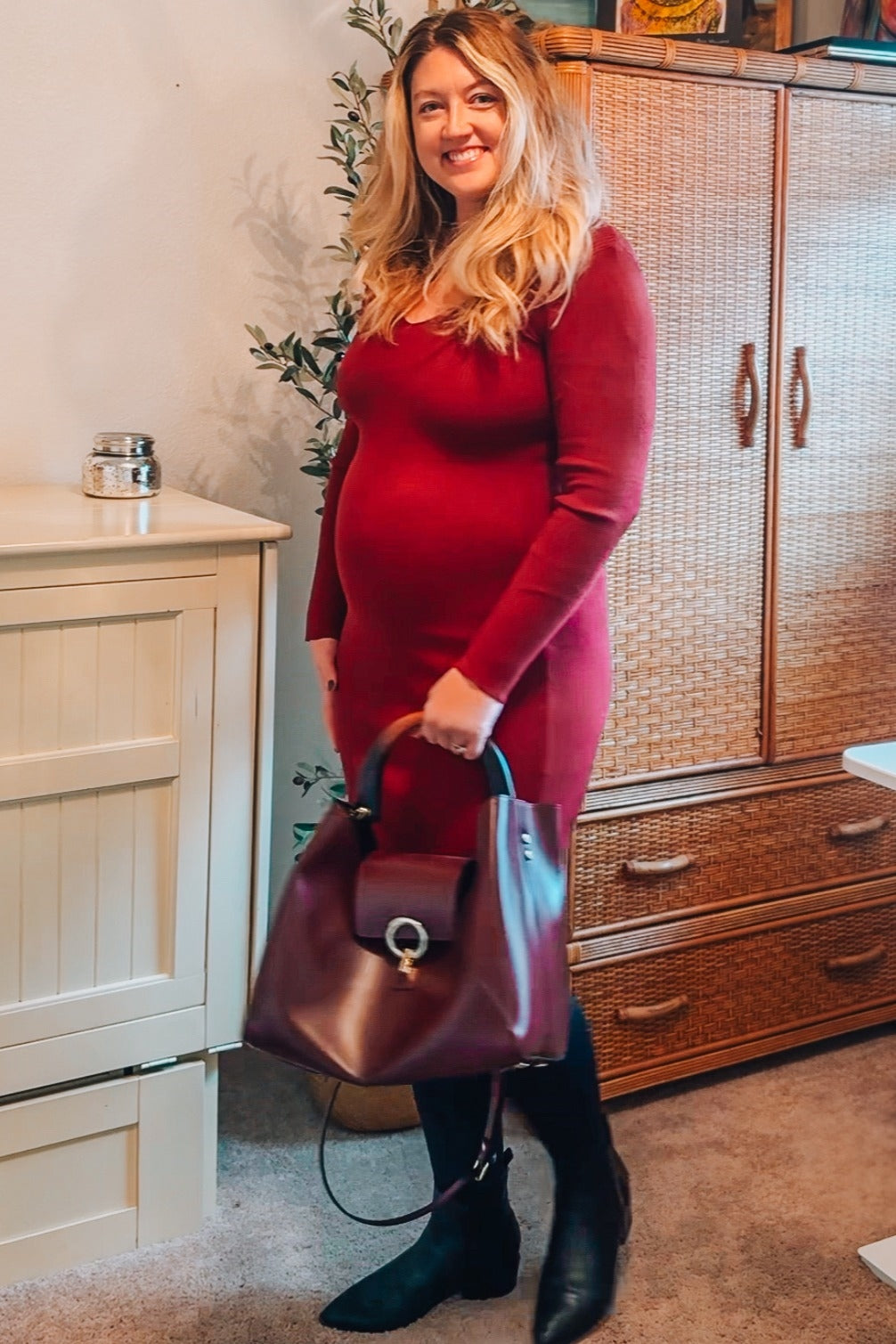 woman wearing a burgundy long sleeve maternity dress, holding a burgundy handbag and showing her baby bump