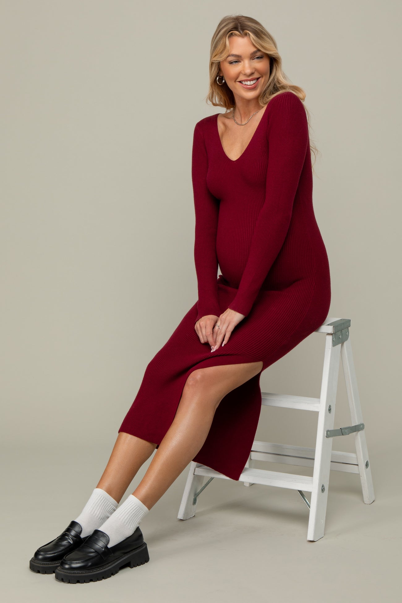 woman wearing a burgundy ribbed maternity dress with a side slit and a visible baby bump, sitting on a white stool