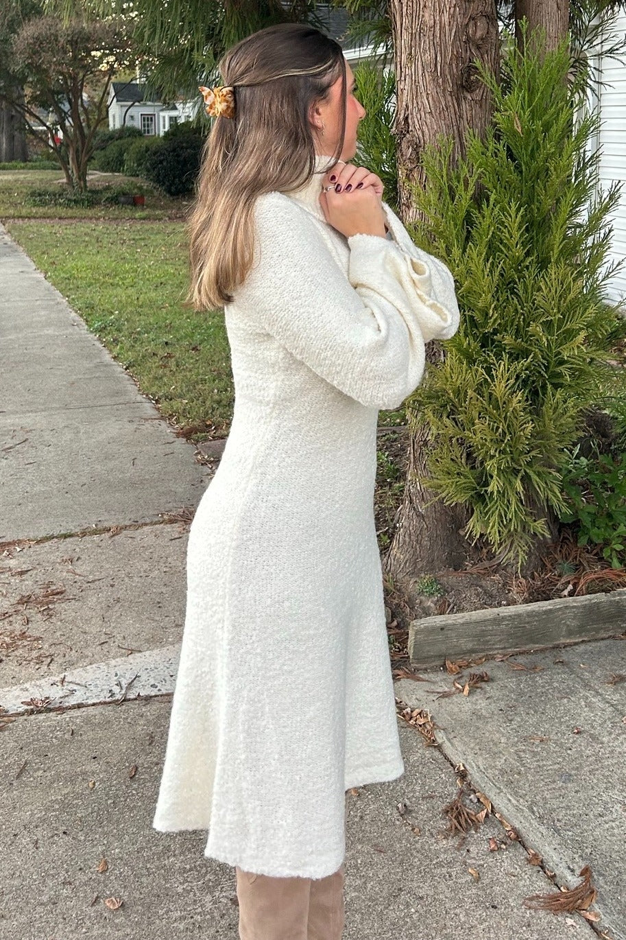 woman wearing a cream-colored long sleeve sweater dress with a flared hem standing outdoors with hands clasped
