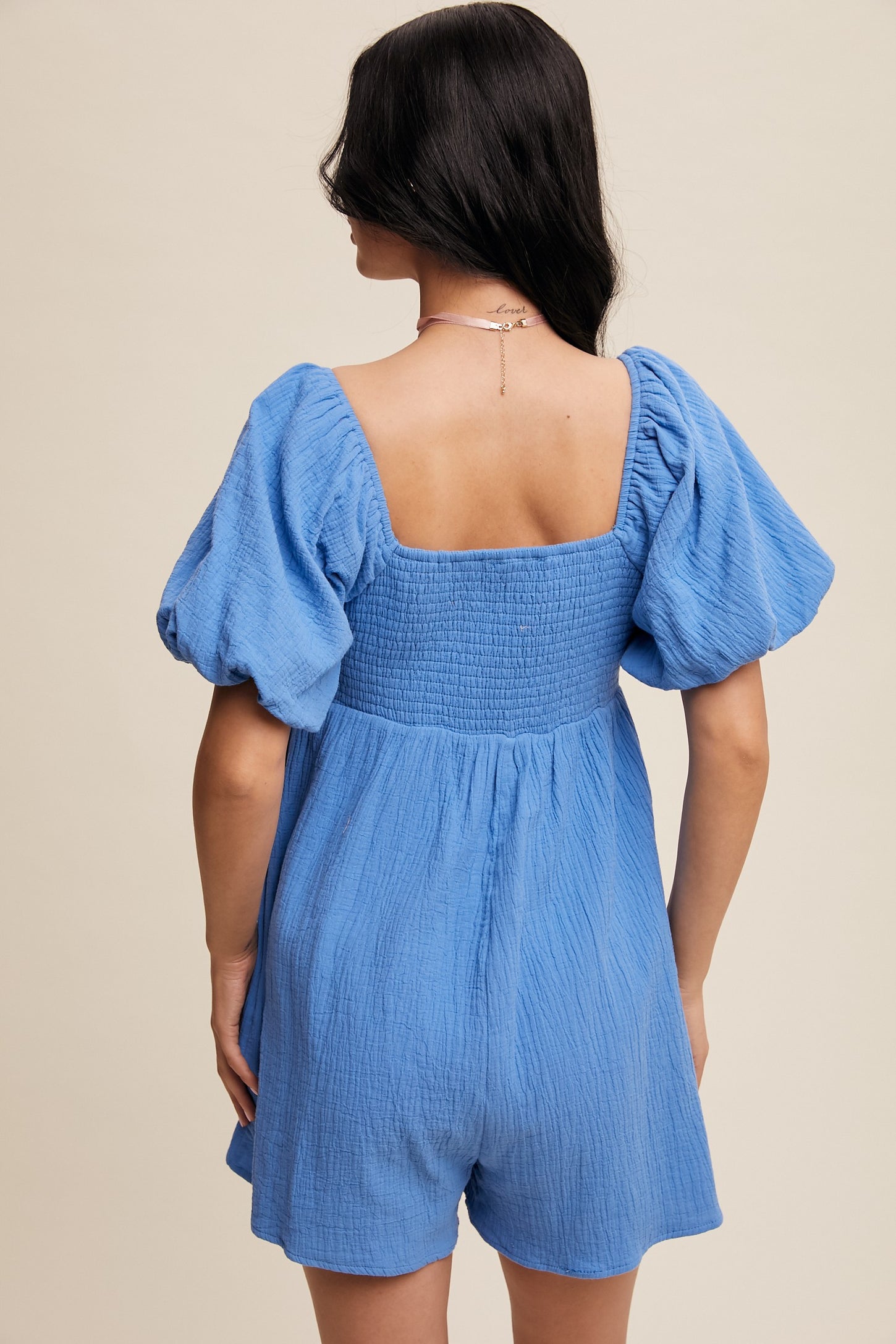 woman wearing a light blue short romper with puff sleeves and a smocked back, seen from the back