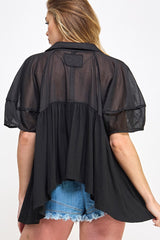 woman wearing a black sheer top with short sleeves and a flowy back, paired with denim shorts