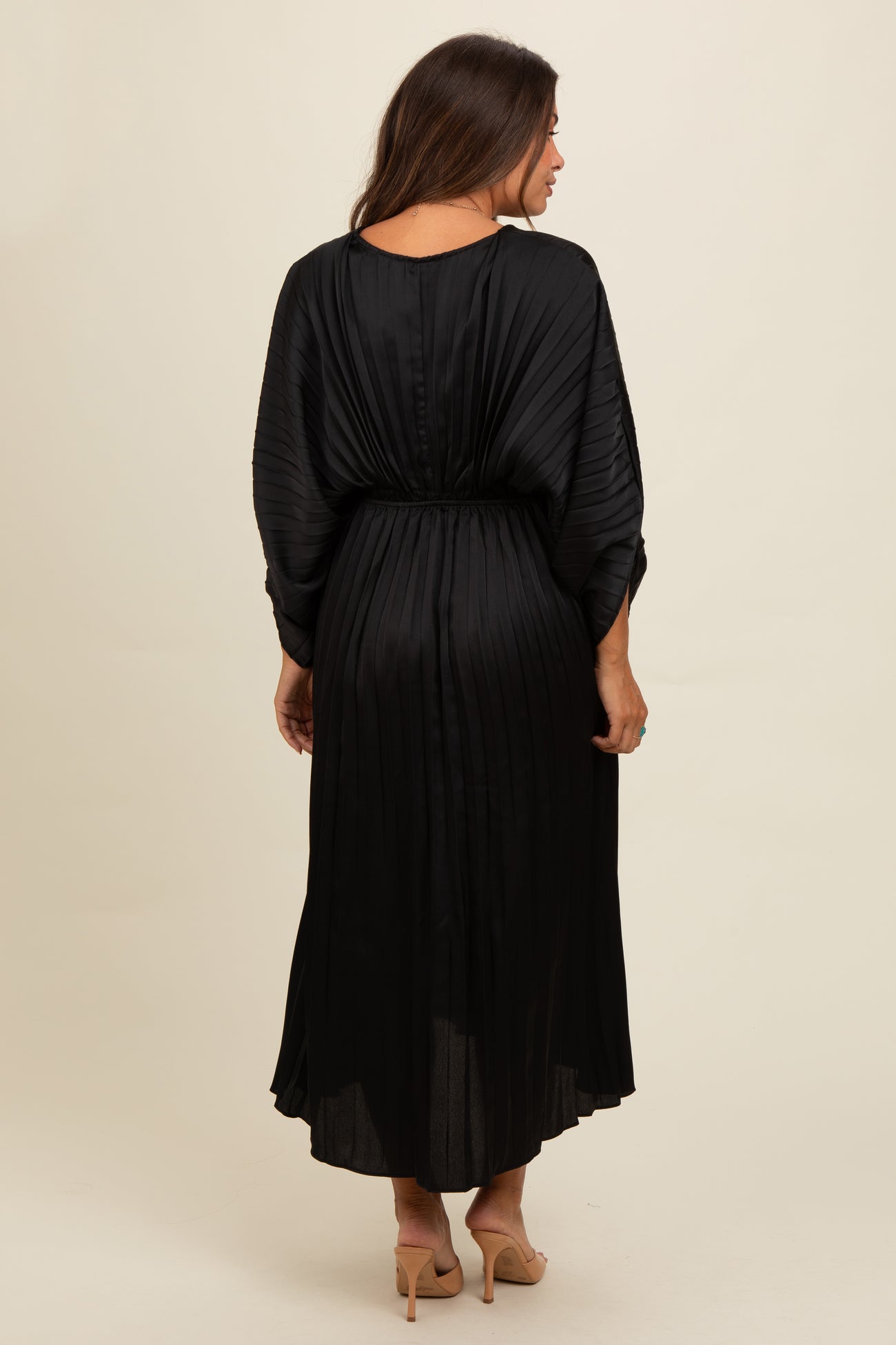 woman wearing a black pleated maternity maxi dress with a cinched waist and long sleeves, showing a baby bump from behind