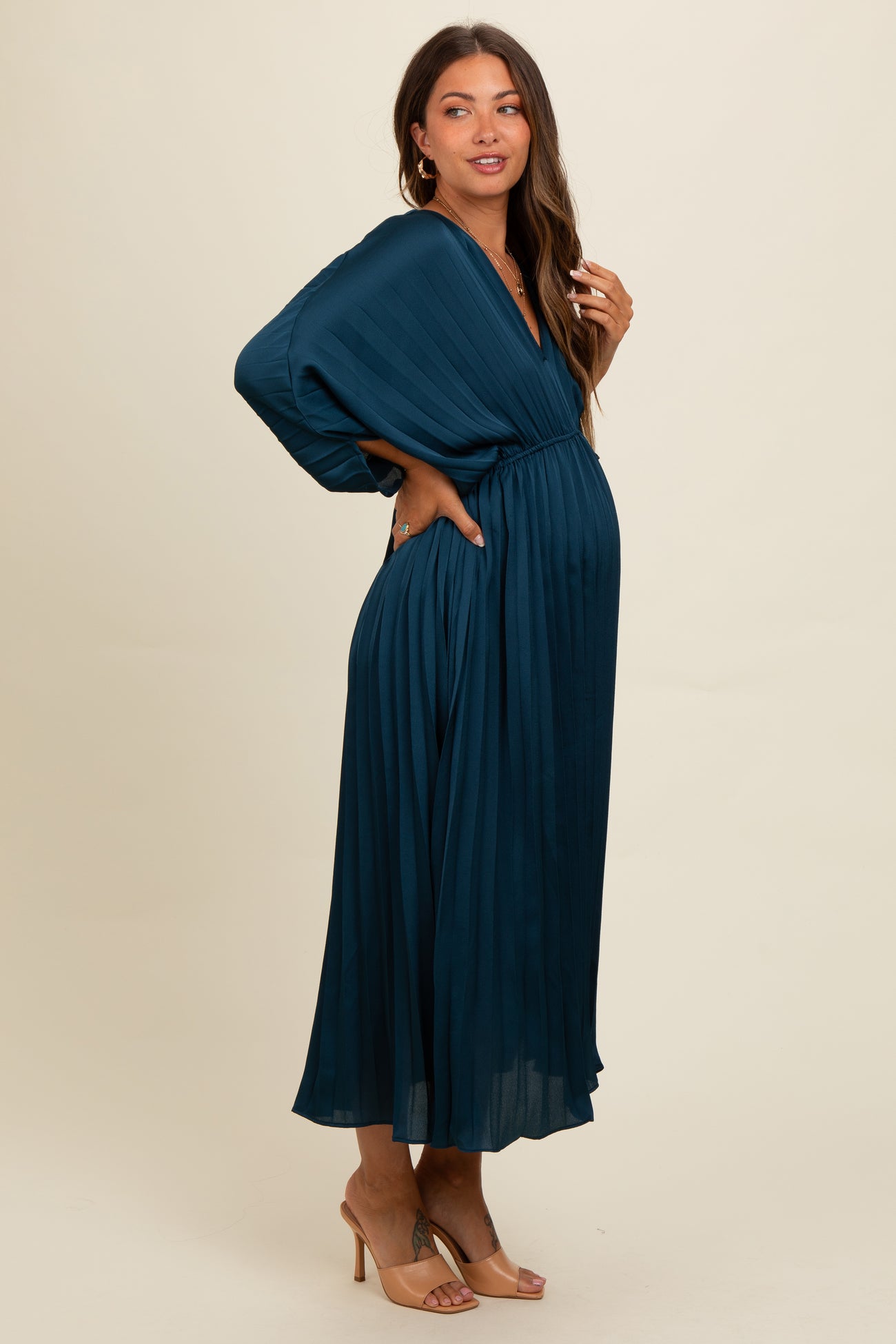 woman wearing a dark teal pleated maternity dress with a deep v-neck and long sleeves, showcasing her baby bump