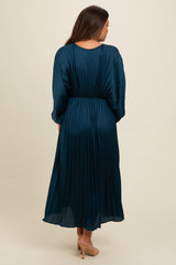 woman wearing a teal pleated maternity dress with three-quarter sleeves and cinched waist, showing a flowing silhouette