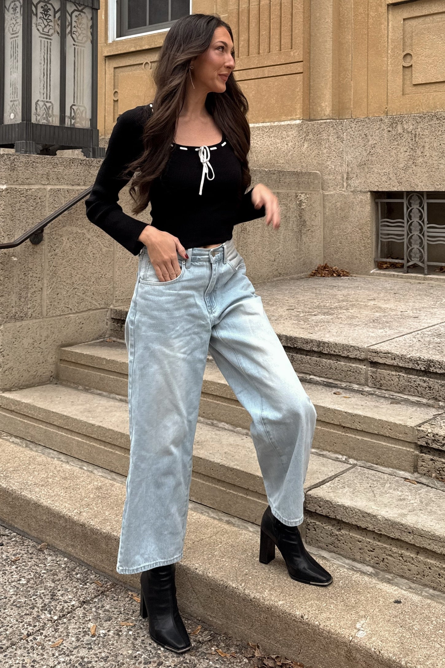 woman wearing a black long sleeve top with tie detail and light wash wide-leg jeans, standing on outdoor steps