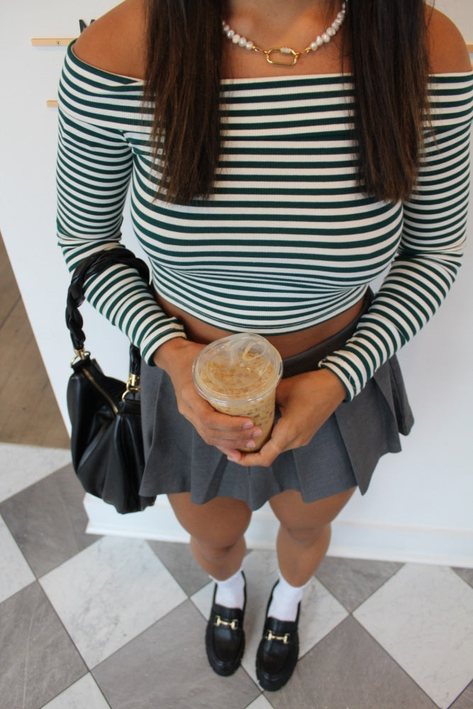 woman wearing a green and white striped off-the-shoulder crop top, gray pleated skirt, holding an iced drink, and black loafers